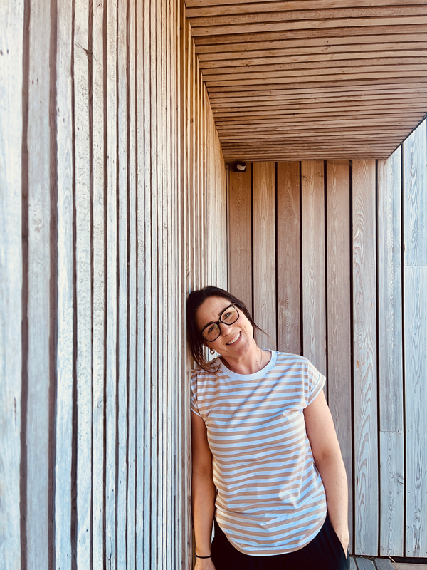Elizabeth McGregor, UGC creator and content writer, full-length in a blue-striped t-shirt leaning against a timber wall
